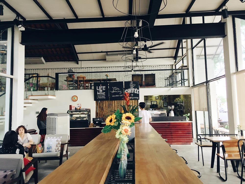 Qu&aacute;n c&agrave; ph&ecirc; với b&agrave;n gỗ d&agrave;i, hoa trang tr&iacute; v&agrave; thực kh&aacute;ch ngồi tr&ograve; chuyện trong kh&ocirc;ng gian hiện đại, s&aacute;ng sủa.