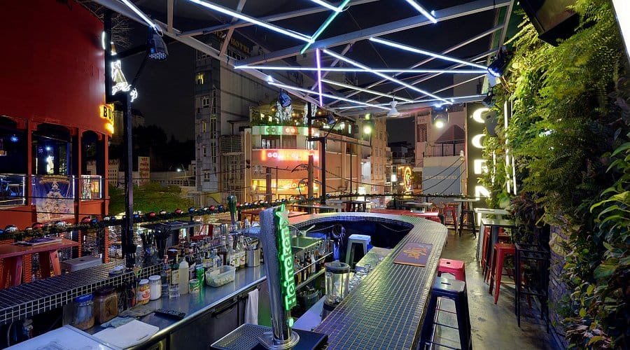 Quán bar sân thượng với ánh sáng neon, quầy bar trung tâm, và bàn ghế xung quanh, không gian về đêm.