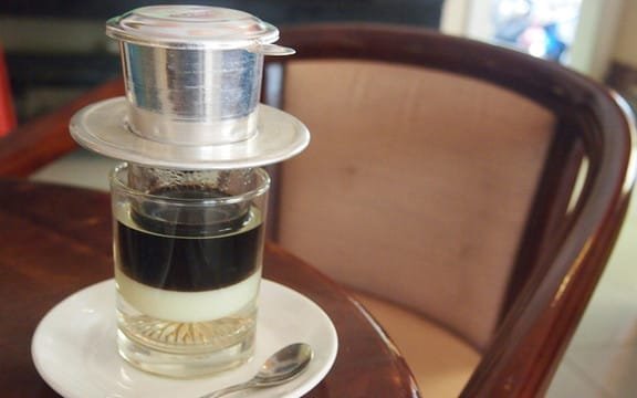 Ly c&agrave; ph&ecirc; sữa đ&aacute; Việt Nam với phin tr&ecirc;n b&agrave;n gỗ, cạnh đ&oacute; l&agrave; muỗng nhỏ v&agrave; ghế tựa.