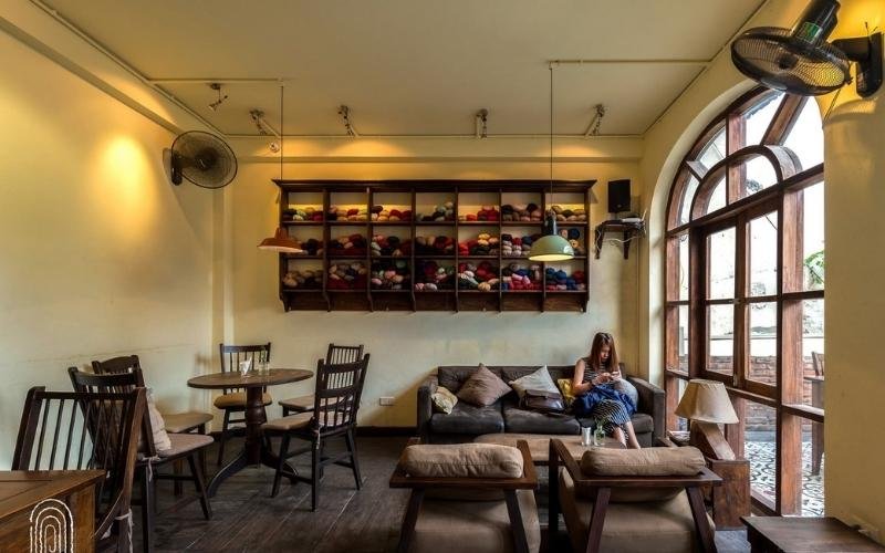 Quán cà phê ấm cúng với bàn ghế gỗ, kệ trưng bày nhiều màu sắc, một người ngồi trên ghế sofa cạnh cửa sổ.
