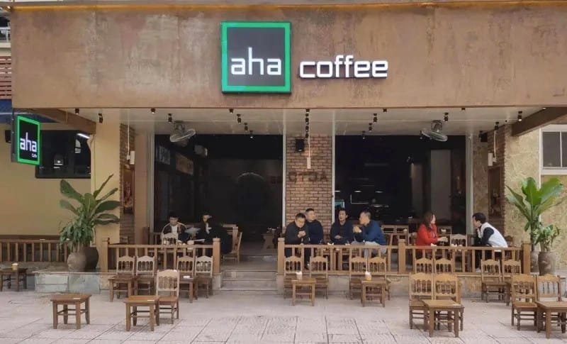 Top 32 thương hiệu nhượng quyền cafe siêu lợi nhuận 10 Bên ngoài một quán cà phê với bảng hiệu "aha coffee", có vài người ngồi trò chuyện trên bàn ghế gỗ.