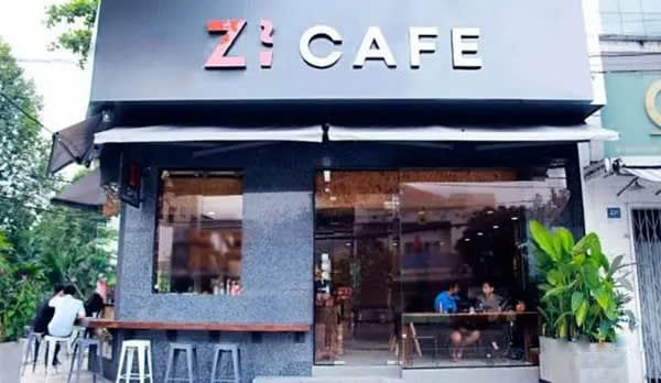 Top 32 thương hiệu nhượng quyền cafe siêu lợi nhuận 28 Mặt tiền quán cà phê hiện đại với cửa kính lớn và cây xanh trang trí bên ngoài, có vài người ngồi bên trong.